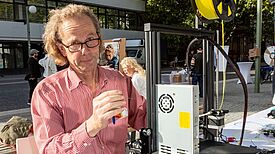 Herr Schellenberg vom Repair Café steht im Freien an einem Tisch mit einem 3D-Drucker und schaut in die Kamera.