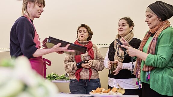 Ein Mitglied von RESTLOS GLÜCKLICH e. V. zeigt drei Frauen die Zubereitung eines Gerichts, das sie bei dem Workshop kennenlernen. 