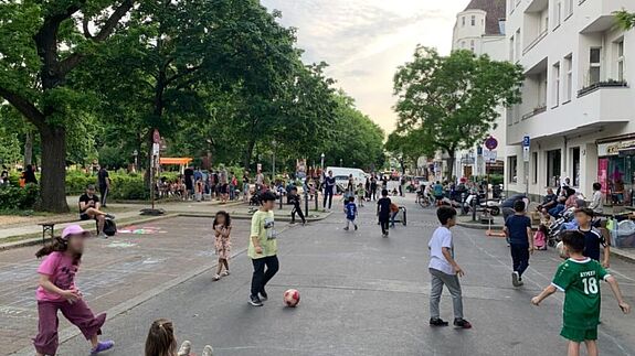 Kinder spielen auf einer temporär gesperrten Straße im Quartier Pankstraße, während Erwachsene die lebendige Atmosphäre genießen.