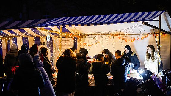 Einige Menschen stehen vor einem erleuchteten Marktstand auf dem Lichtermarkt im QM-Gebiet Klixstraße / Auguste-Viktoria-Allee.