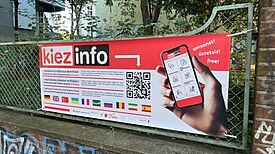 Ein Banner der „kiezinfo“-App mit QR-Code und mehrsprachiger Beschreibung hängt an einem Zaun im QM-Gebiet Zentrum Kreuzberg.