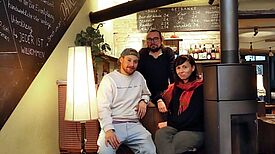 Zwei Engagierte sitzen an der Bar im Kieztreff „Kamine&Wein“, eine weitere Person steht hinter ihnen. 