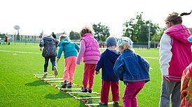 Kinder spielen gemeinsam auf einem Sportplatz und üben eine Koordinationsübung auf einer Balancierleiter.